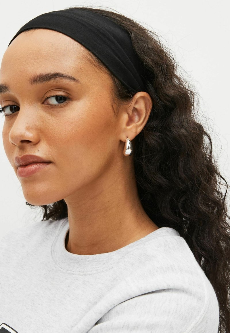 Sølv dråbeørering med blank finish, båret i venstre øre. Sort stofhovedbånd med glat tekstur. Grå sweat-shirt med rund hals.