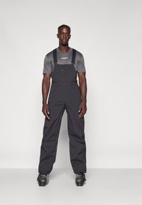 Svarte isolerte bib-overaller med justerbare stropper, glidelås frontlomme og elastiske cuff-er. Båret over en tettsittende grå T-skjorte med korte ermer.