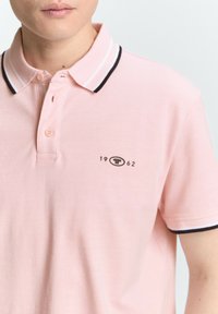Polohemd in hellrosa mit einem klassischen Kragen mit schwarz-weißen Streifen, einer Zwei-Knopf-Leiste und einem Logo mit "1962" auf der Brust.