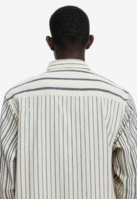 Camisa a rayas con líneas verticales en negro y crema. La tela parece ligera, con un cuello clásico y detalles de botones en la parte posterior.