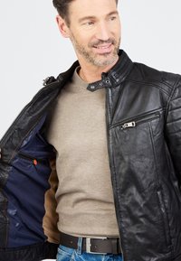 Schwarze Leder-Motorradjacke mit Reißverschluss, Druckknöpfen und Innentaschen. Getragen über einem beigen Hemd und blauen Jeans.