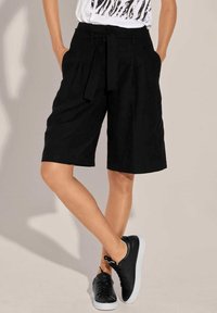 Black linen shorts with a tied waistband, featuring side pockets, a relaxed fit, and knee-length cut, paired with black sneakers.