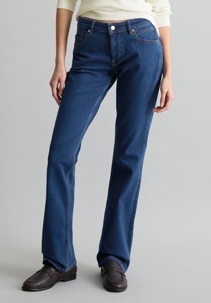 Femme portant un jean droit bleu et des mocassins marron foncé, debout devant un fond gris clair uni.