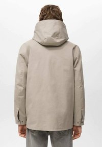 Mężczyzna ubrany w beżową kurtkę z kapturem i szare dżinsy, widziany od tyłu na jednolitym, jasnym tle.