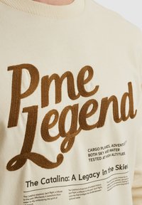 Sudadera color crema con gran texto bordado en marrón "Pme Legend" y detalles adicionales impresos sobre el hidroavión Catalina.