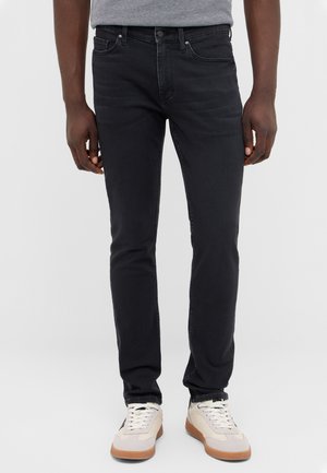 Male model wearing black slim-fit jeans and white sneakers with gum soles, standing against a plain light gray background.