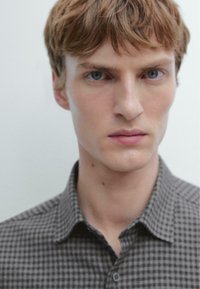 Närbild på en manlig modell som bär en grå rutig skjorta med krage. Tyget har en jämn struktur och pryds av små mörka fyrkanter.