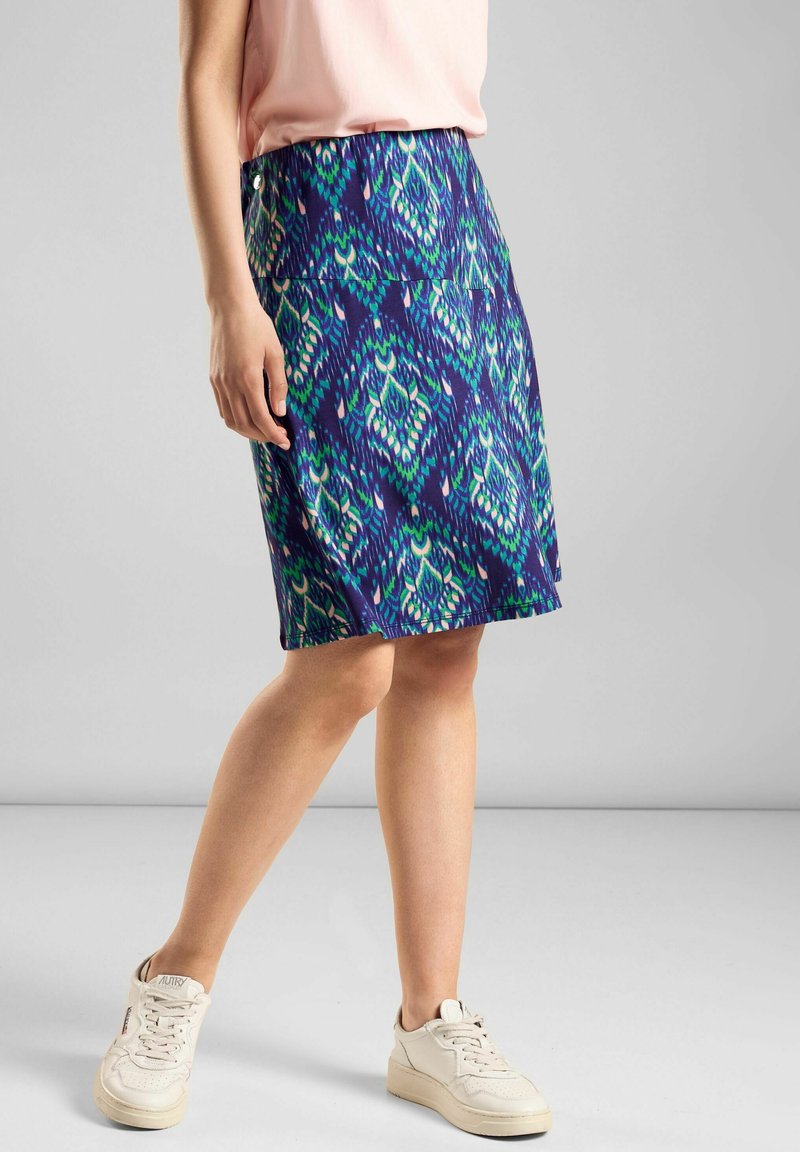 Femme portant une jupe à motifs bleus et verts longueur genou, un haut rose clair et des baskets blanches, debout devant un fond gris uni.