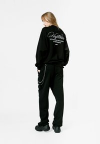Sudadera negra de gran tamaño con texto blanco en la parte posterior, combinada con pantalones negros que tienen un accesorio de cadena de metal. El modelo está de espaldas.