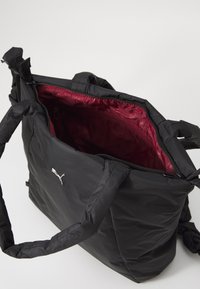 Zwarte tote bag van zacht, matte stof. De binnenzijde is gevoerd met bordeauxrood en heeft een ritssluiting. Heeft een klein zilveren logo aan de buitenkant.