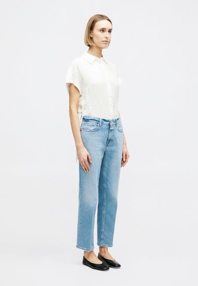 Femme debout, légèrement tournée vers la droite, portant une chemise blanche à manches courtes boutonnée, un jean bleu clair et des chaussures plates noires sur fond blanc.