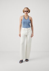 Top de mezclilla tipo halter, pantalones anchos blancos y sandalias de tacón azules con detalle de nudo. La modelo lleva gafas de sol oscuras, mostrando un atuendo casual.
