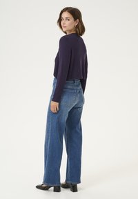 Maglione viola a maniche lunghe accorciato abbinato a jeans blu a vita alta con gamba larga e scarpe con tacco a blocco nere. Vista posteriore.