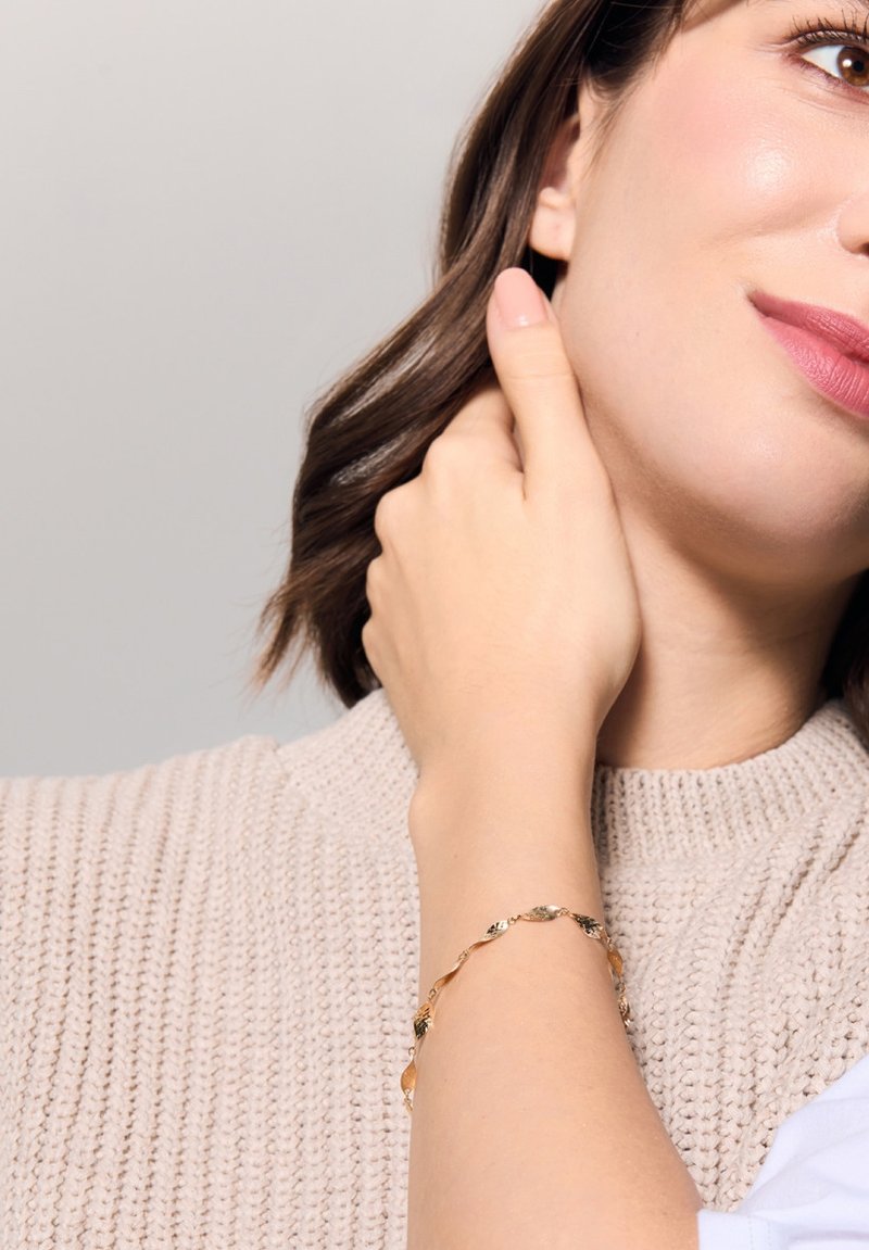 Złota bransoletka z falistymi ogniwami i akcentami w kształcie liści, noszona na nadgarstku na tle teksturowanego beżowego swetra. Gładkie, błyszczące wykończenie.