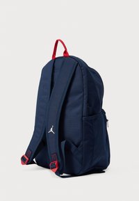 Σακίδιο πλάτης σε ναυτικό μπλε με κόκκινες λεπτομέρειες, υφασμάτινο υλικό, επενδεδυμένα σχήματα, ρυθμιζόμενοι ιμάντες, πλαϊνές τσέπες και λευκό λογότυπο στο μπροστινό μέρος.