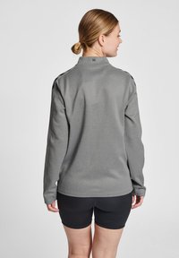Grå, langærmet pullover med høj krave, der har struktureret stof og sorte detaljer på skuldrene, iført over sorte shorts.
