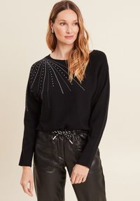 Schwarzer Pullover mit langen Ärmeln und Rundhalsausschnitt, verziert mit metallic Perlenverzierungen, die ein strahlendes Muster bilden. Kombiniert mit schwarzen Lederhosen.