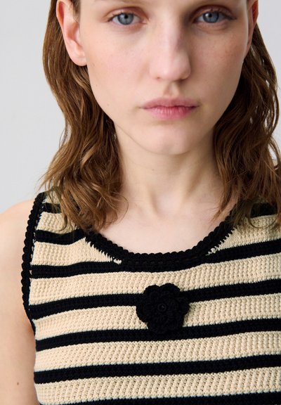 Jersey de punto con rayas horizontales en negro y beige, cuello redondo y un adorno de flor de ganchillo negro en el centro.