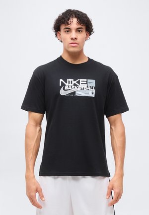 Young man with curly hair wearing a black Nike Basketball T-shirt and white sports shorts standing against a plain background.