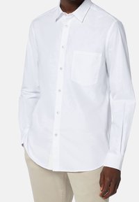 En person som bär en välsittande vit skjorta med knäppning och bröstficka, stående mot en enfärgad bakgrund.