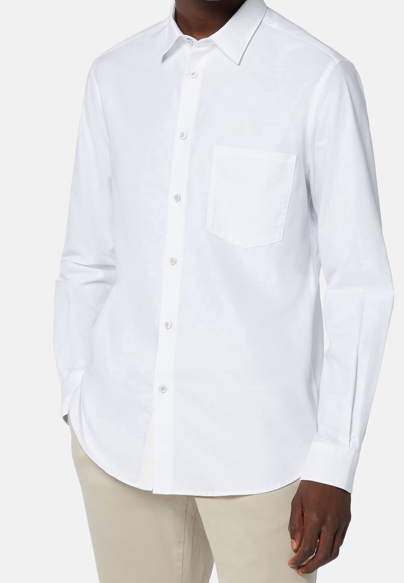 En person som bär en välsittande vit skjorta med knäppning och bröstficka, stående mot en enfärgad bakgrund.