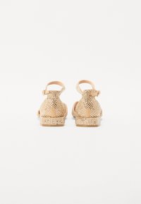 Beige sandaalit, joissa on teksturoitu päällinen, metalliset niitit ja solkikiinnitys. Littee korko ja avoin muoto takana.