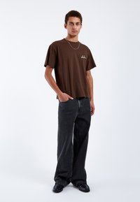 Brown cotton t-shirt with short sleeves and white embroidered detailing. Paired with loose-fit black denim jeans and black shoes.