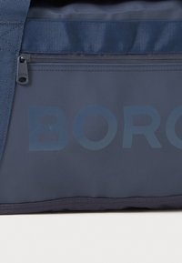 Bolsa de deporte azul marino con una textura suave, que presenta un cierre de cremallera, un logo destacado de "BORG" y una correa de hombro trenzada.
