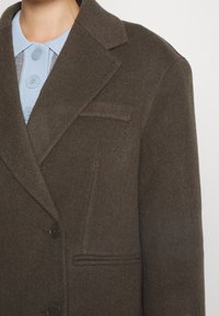 Gros plan d'une personne portant un blazer en laine marron foncé sur une chemise bleu clair à col avec trois grands boutons.