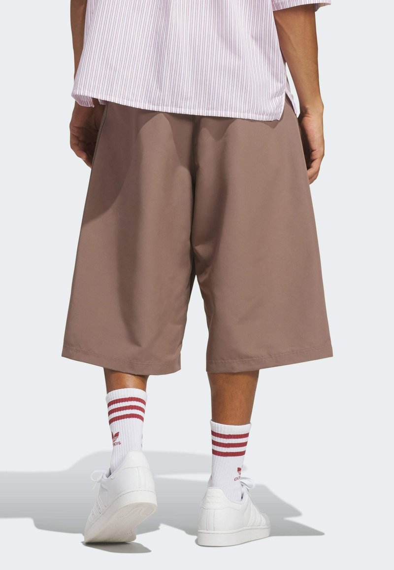 Person wearing loose brown knee-length shorts, white sneakers, and white socks with red stripes standing against a plain background.