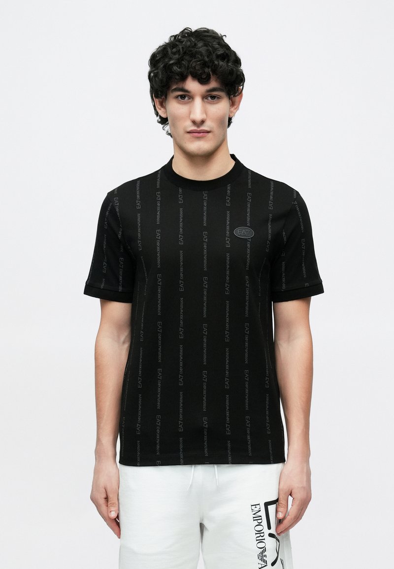 Fiatal férfi göndör hajjal, fekete EA7 Emporio Armani pólót és fehér Emporio Armani logós rövidnadrágot viselve.