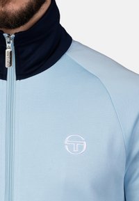 Lichtblauwe jas met een marineblauwe kraag, voorrits en een witte geborduurde logo. De stof lijkt glad met een getailleerd ontwerp.