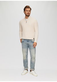 Pull en maille beige clair à demi-zip avec un motif texturé, associé à un jean bleu clair déchiré et des baskets blanches.
