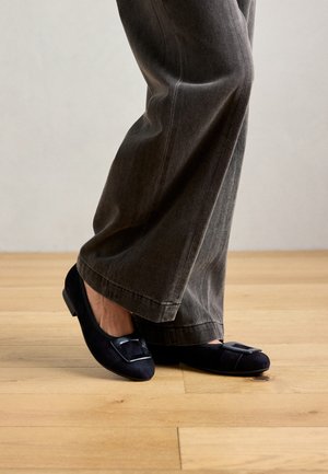 Persona vestida con pantalones sueltos grises y suaves bailarinas de ante negro con hebillas decorativas cuadradas, de pie sobre un suelo de madera clara.