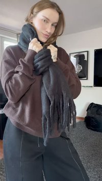 Noor naine toas, seljas pruun sviiter ja mustad püksid, reguleerib paksu tumedashallide salli oma kaela ümber.