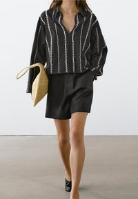 Musta kauluspaita valkoisilla pystysaumoilla, yhdistetty mustiin shortseihin ja palmikkokuituiseen vaaleaan laukkuun. Sileä kangas rentoa istuvuutta.