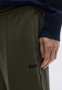 Olívazöld futónadrág texturált anyagból, oldalsó csíkkal és a "BOSS" logóval fekete színben a zseb közelében, kéz a derékpánton pihen.
