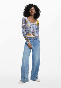 Áttetsző hosszú ujjú felső absztrakt kék és sárga mintával, laza szabású világoskék farmernadrággal, amely egyedi varrási részletekkel rendelkezik.