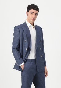 Dobbeltspent marineblå blazer med metallknapper, laget av teksturert stoff. Kombinert med en hvit skjorte med krage og matchende bukser.