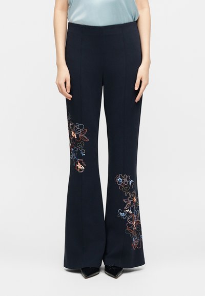 Μπλε σκούρο παντελόνι με φαρδιά μπατζάκια και πολύχρωμη κεντημένη λουλουδένια διακόσμηση μπροστά στους μηρούς και στα κάτω μέρη των ποδιών, φορεμένο με ανοιχτό μπλε τοπ και μαύρα μυτερά παπούτσια.