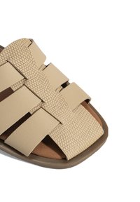 Beige gevlochten sandalen met ingeweven banden, textuurafwerking, vierkante teenvorm en een bruine binnenzool op een lichtbruine buitenzool.