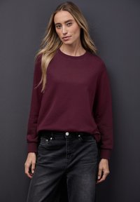 Sudadera burdeos con cuello redondo y mangas largas, combinada con jeans negros con cierre de botones y un leve desgaste.