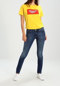 Žluté tričko s červeným potiskem "HIGHWAY HONEY" v kombinaci s tmavě modrými úzkými džíny a bílými teniskami. Klasický Outfit s jednoduchým designem.