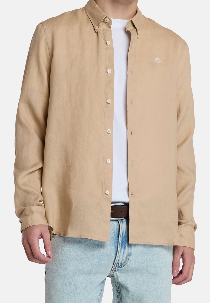 Homme portant une chemise beige boutonnée sur un t-shirt blanc, associée à un jean bleu clair et une ceinture marron, visible du cou jusqu'aux hanches.