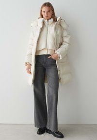 Voering, crèmekleurige jas met een hoge kraag en ritssluiting. Gelaagd over een fleece-achtige, kort- mouwtop. Gecombineerd met grijze jeans met wijde pijpen.