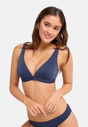 Młoda kobieta z długimi brązowymi włosami, ubrana w granatowe bikini, uśmiecha się i lekko patrzy na bok na gładkim tle.