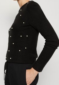 Svart stickad tröja med rund halsringning, med utspridda vita pärldetaljer på framsidan och slim-fit långärmar.