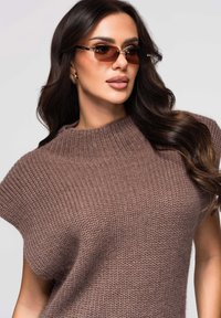 Pull en tricot marron à col montant, avec un motif texturé. Le mannequin porte des lunettes de soleil teintées et a les cheveux longs et ondulés.
