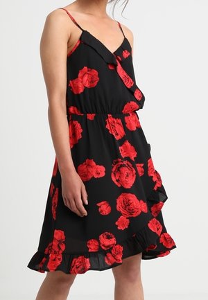 Kvinde iført en sort ærmeløs kjole med rødt blomsterprint og flæsekant, stående mod en ensfarvet lys baggrund.