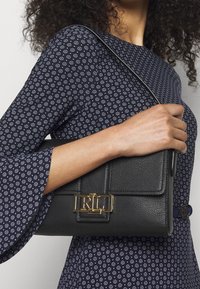 Svart läderhandväska med texturerad yta, guldfärgad låsdetalj med "RL" och en smal axelrem. Har en strukturerad design.